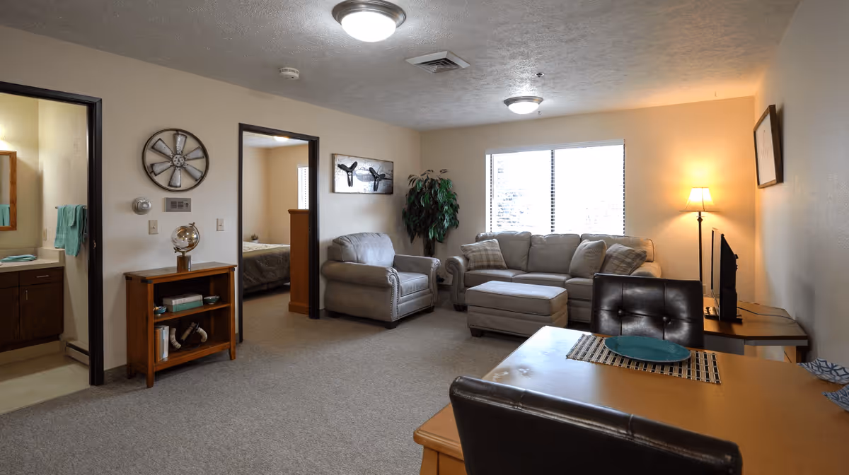 A cozy living room area in a retirement living facility with a beige armchair, a matching sofa with plaid pillows, and an ottoman. There is a large window with blinds letting in natural light, a floor lamp in the corner, and a small table with a TV. Adjacent to the living room is a dining table with two black leather chairs and a blue plate on a placemat. To the left, there is an open doorway leading to a bathroom with a sink and towels, and another doorway leading to a bedroom with a bed and wooden headboard. The walls are light beige, and there is a small wooden bookshelf with decorative items and a globe. The ceiling has two round light fixtures.