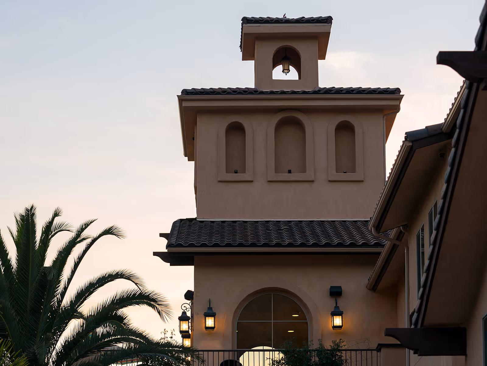 Exterior view of a building with a tower-like structure featuring arched windows and a small bell with a lantern hanging inside. The building has a tiled roof and is partially framed by palm tree leaves on the left side. The sky is clear with a soft evening light.
