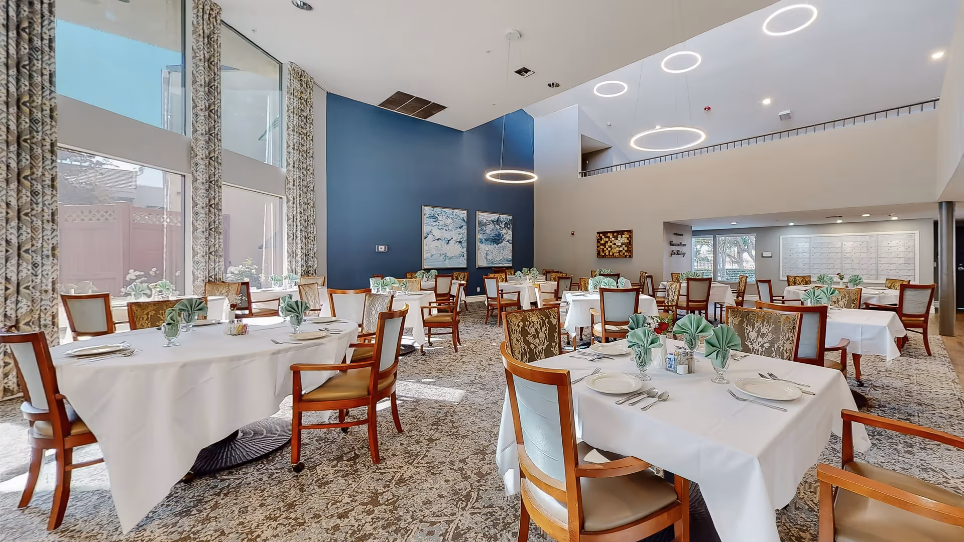 A bright and spacious dining room with multiple tables covered in white tablecloths, each set with plates, silverware, and green folded napkins. The room features large windows with patterned curtains allowing natural light to fill the space, modern circular ceiling lights, and a blue accent wall with artwork. Wooden chairs with cushioned seats surround the tables.