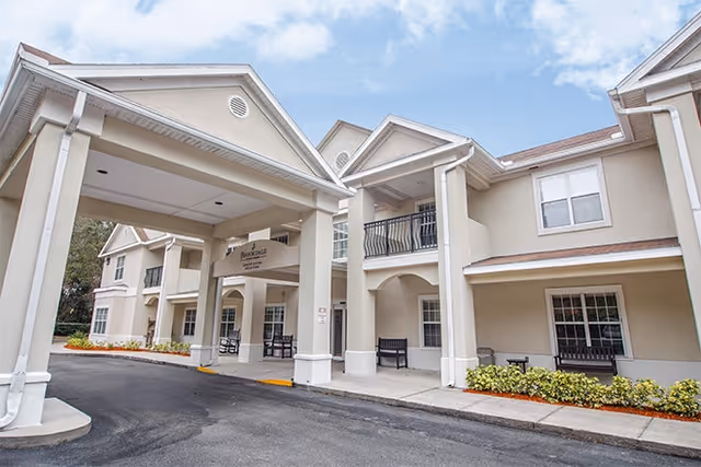 Exterior view of the Brookdale Wekiwa Springs senior living facility showing a beige two-story building with a covered entrance, balconies, windows, and benches along the sidewalk. The sky is partly cloudy.