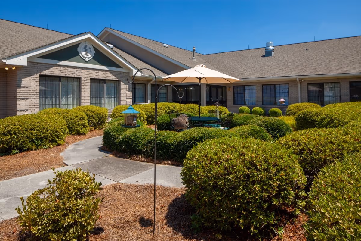 Outdoor courtyard area of a senior living facility with neatly trimmed bushes, a concrete pathway, and a patio table with an umbrella. The building has large windows and a sloped roof under a clear blue sky.