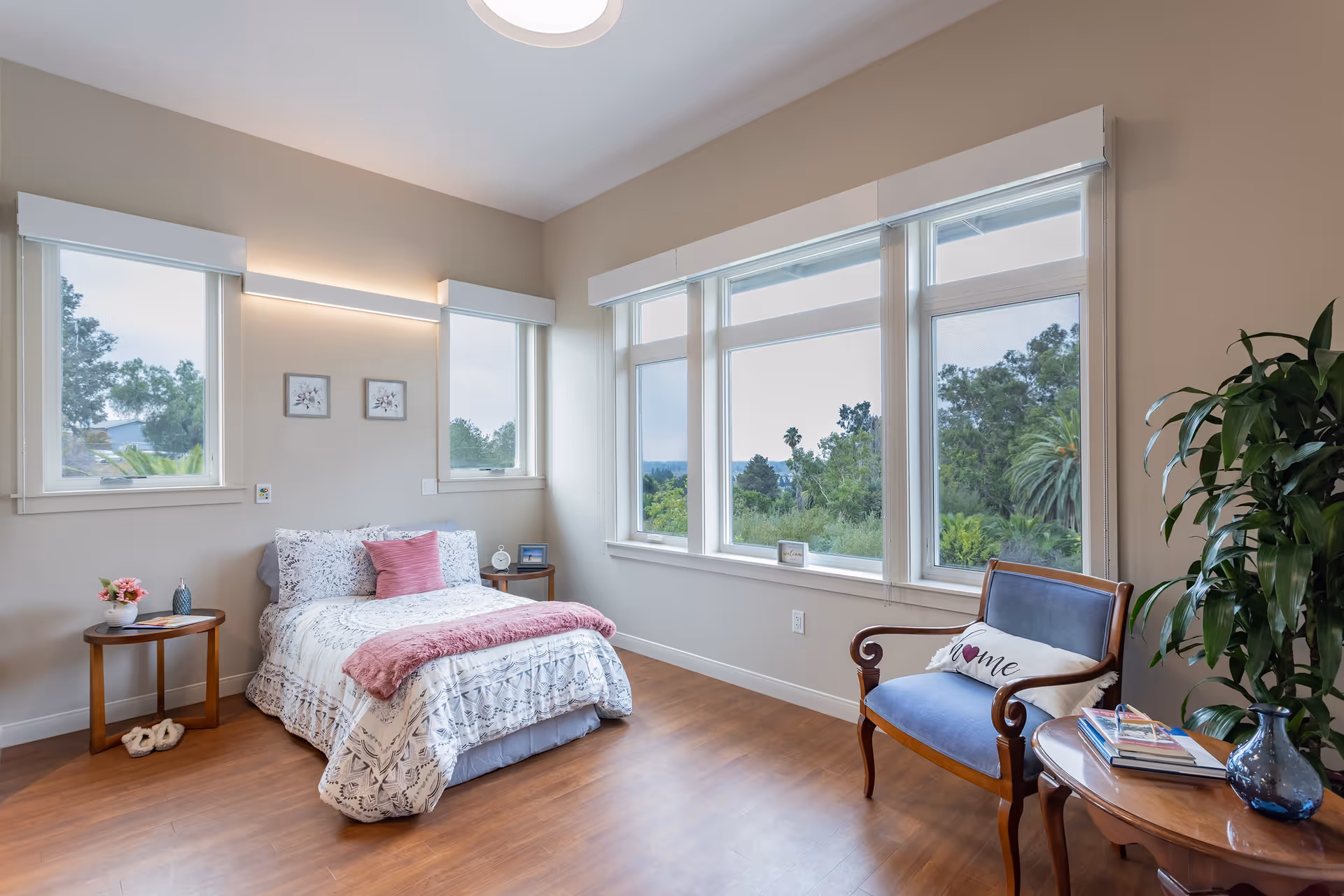 A bright and cozy bedroom with a single bed dressed in white and pink bedding, two small round wooden side tables with decorative items, a wooden chair with a blue cushion and a 'home' pillow, a large plant, and multiple windows showing a green outdoor view.