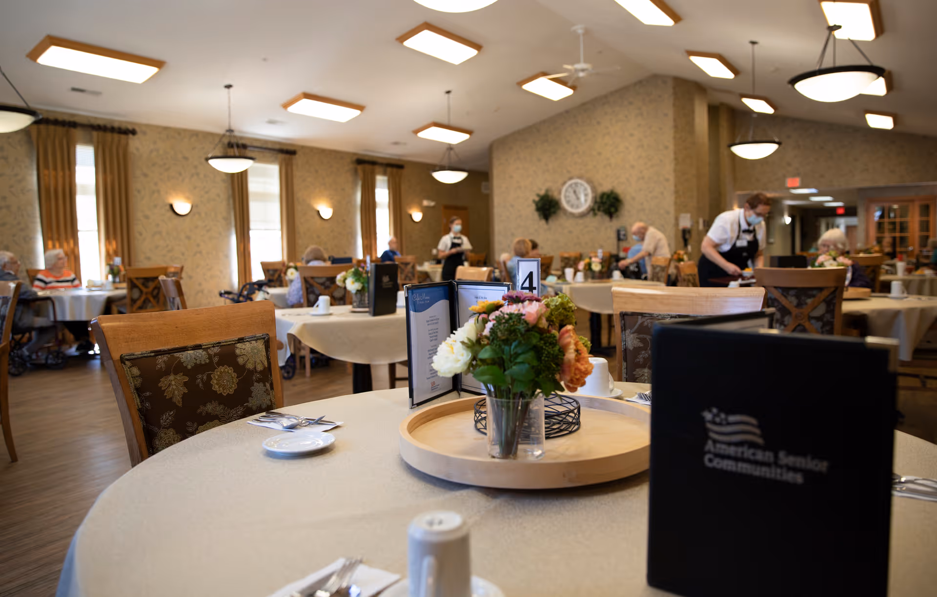 Well-lit assisted living dining room with round tables, floral centerpieces, menus, and staff attending residents.