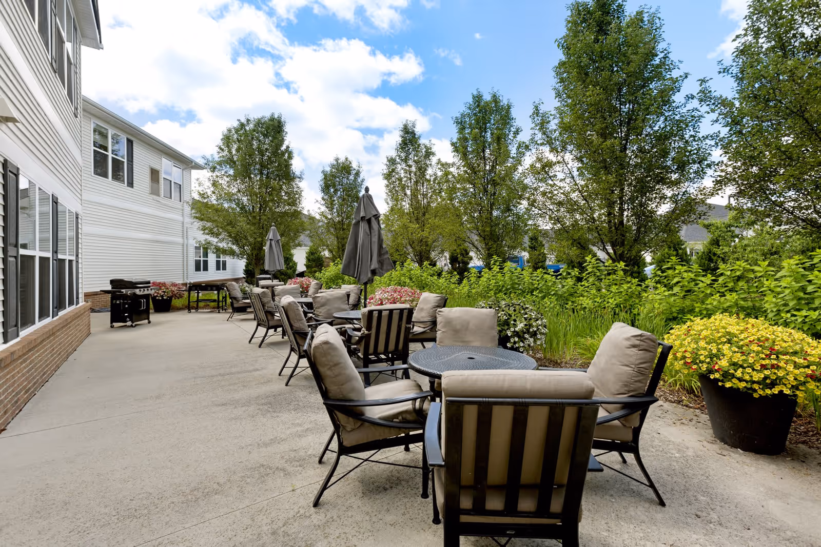 Outdoor patio area at Olivia's Assisted Living with several cushioned chairs and round tables, some with closed umbrellas. The patio is adjacent to a building with large windows and surrounded by green trees and flowering plants under a partly cloudy sky.