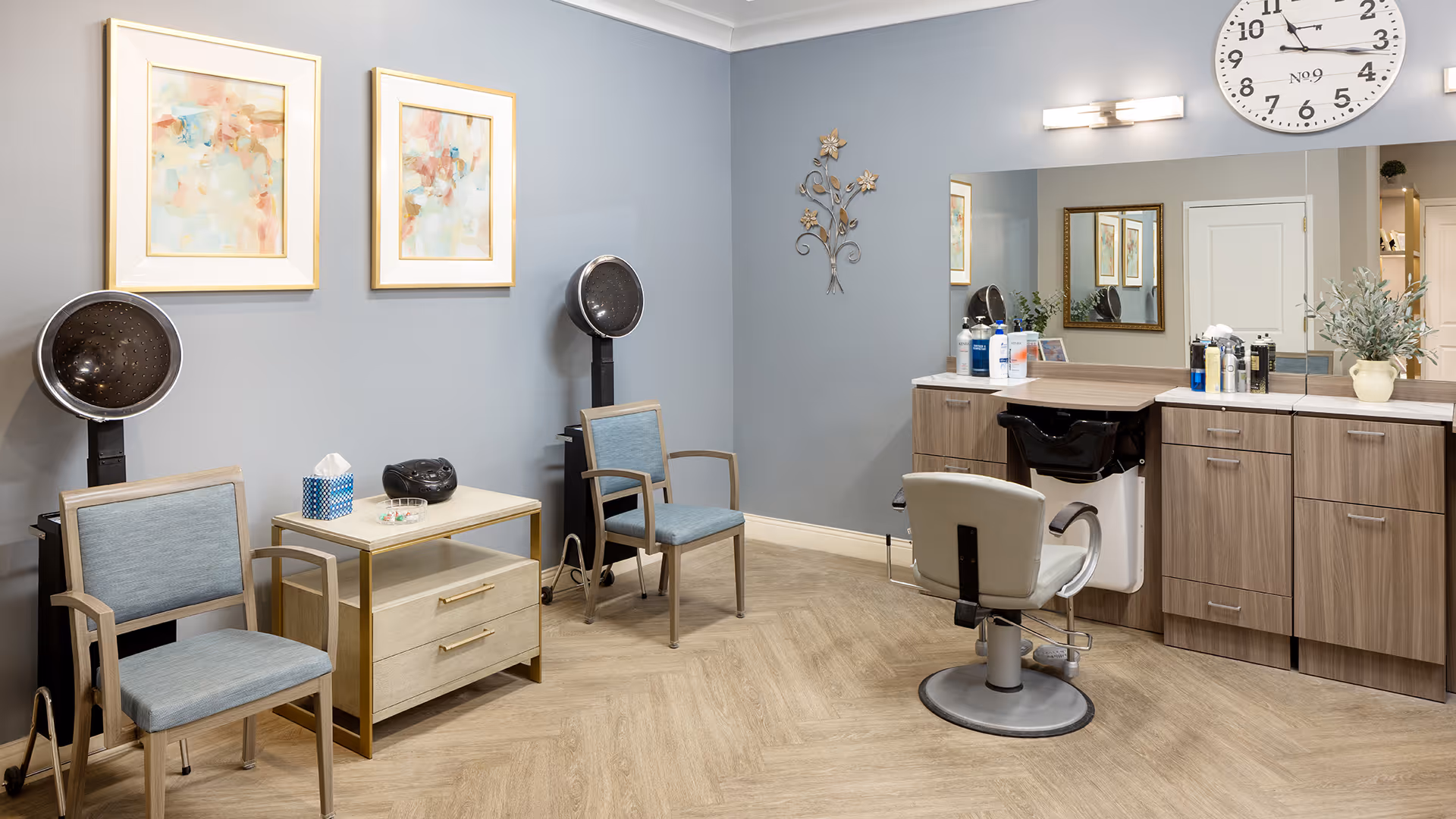 Interior view of a salon area in a senior living facility with two hair drying chairs, a small table with tissue box and radio, a styling chair in front of a large mirror, and wooden cabinets. The walls are painted light blue with two abstract paintings and a decorative metal wall art.
