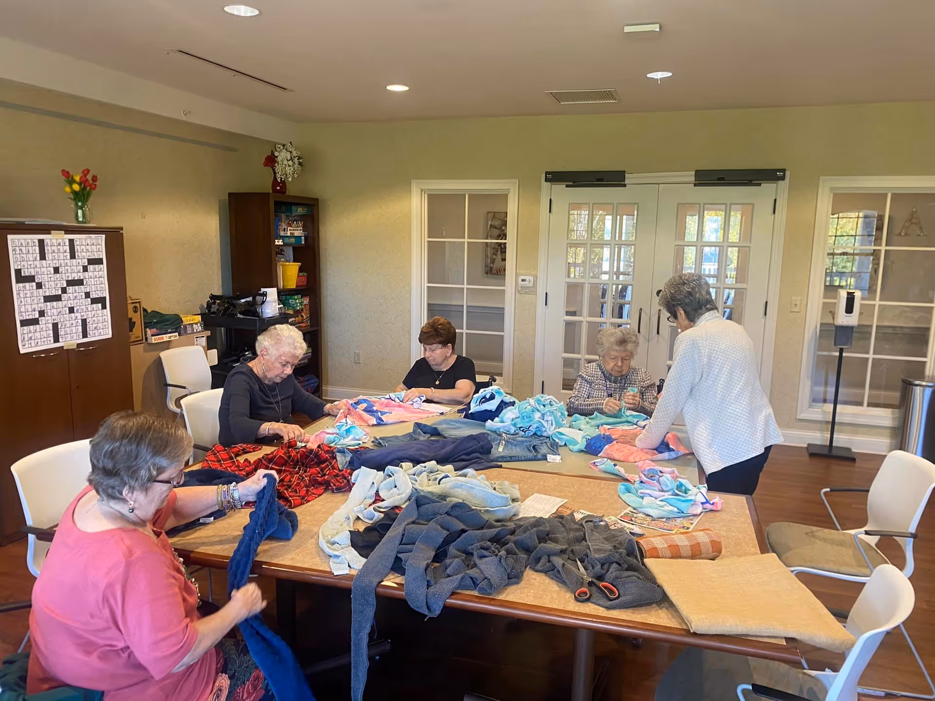 A group of elderly women sitting and standing around a large table in a well-lit room, working on folding and organizing colorful fabric pieces. The room has light-colored walls, wooden floors, and glass-paneled double doors in the background. There is a crossword puzzle on a board on the left wall and shelves with games and supplies.