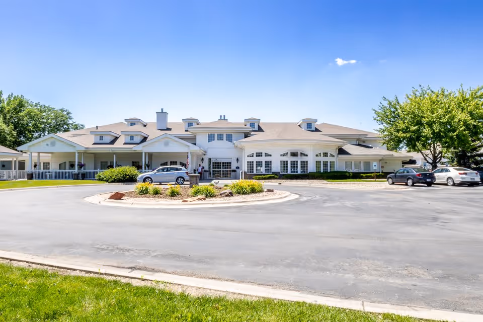 Front exterior view of Bailey Pointe at Roxbury Park, a single-story building with multiple dormer windows, a covered entrance, and a circular driveway with parked cars. The sky is clear and blue, and there are trees and landscaping around the building.