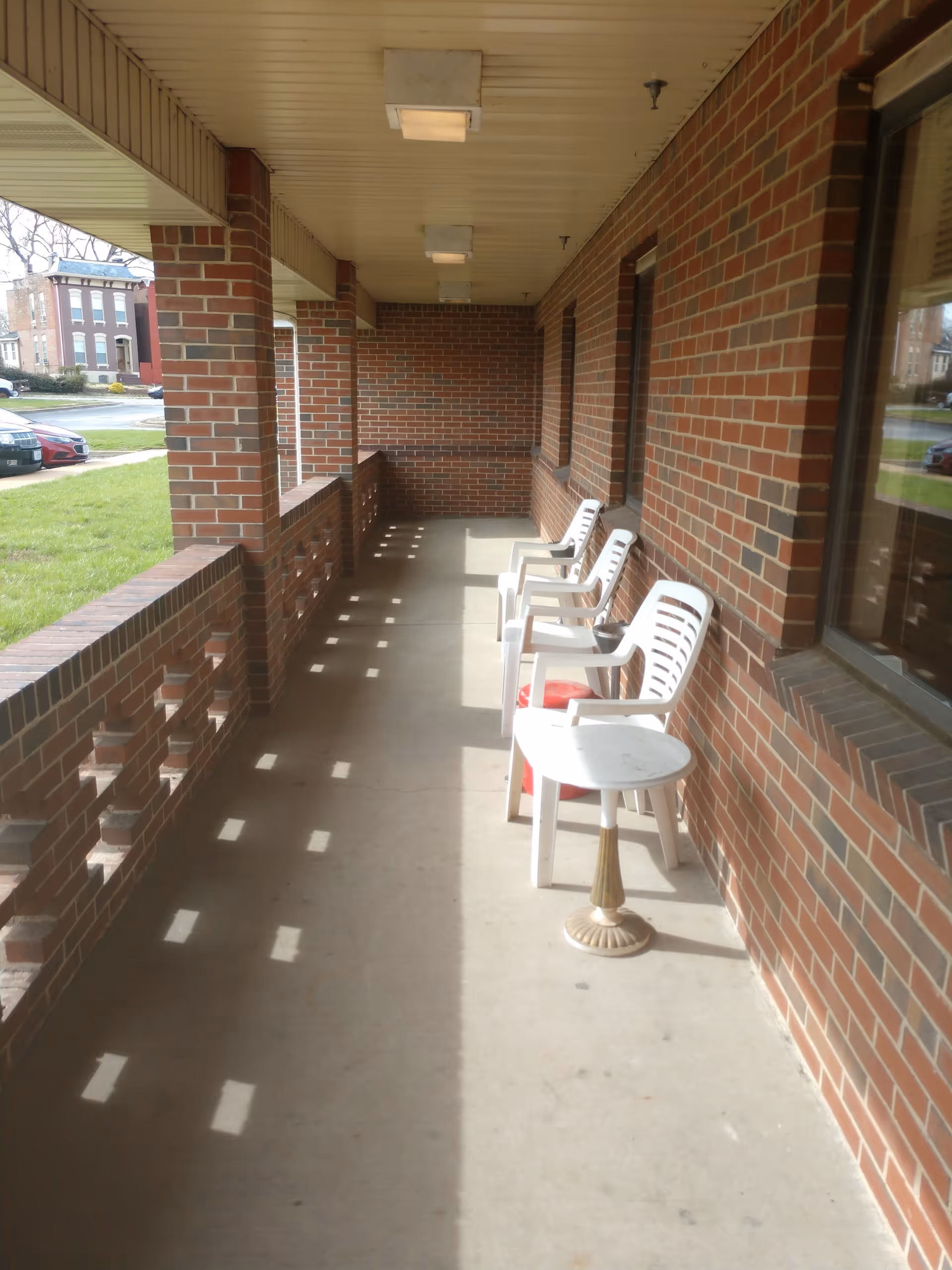 Covered brick porch with a row of white plastic chairs and a small table along a building exterior.
