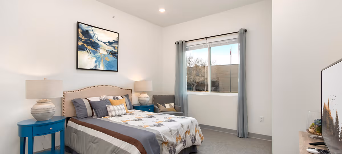 A neatly made bed with a patterned quilt and multiple pillows in a bright bedroom. There are two blue nightstands with matching lamps on either side of the bed. A modern abstract painting hangs above the headboard. A window with light blue curtains lets in natural light, and a gray armchair is positioned near the window. A flat-screen TV is visible on the right side of the image.