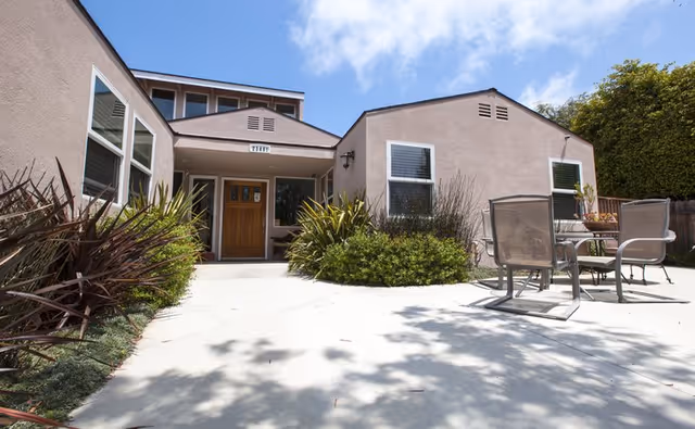 Exterior view of a single-story building with beige walls and a wooden front door, surrounded by plants and shrubs. There is a concrete patio area with a glass-top table and four chairs under a partly cloudy blue sky.