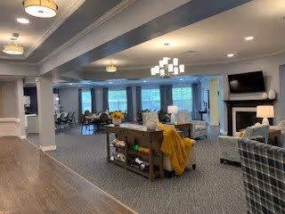 Spacious senior living common area with comfortable seating including armchairs and sofas, a fireplace with a mounted TV above it, a central table with decorative items, and large windows with blue curtains letting in natural light.