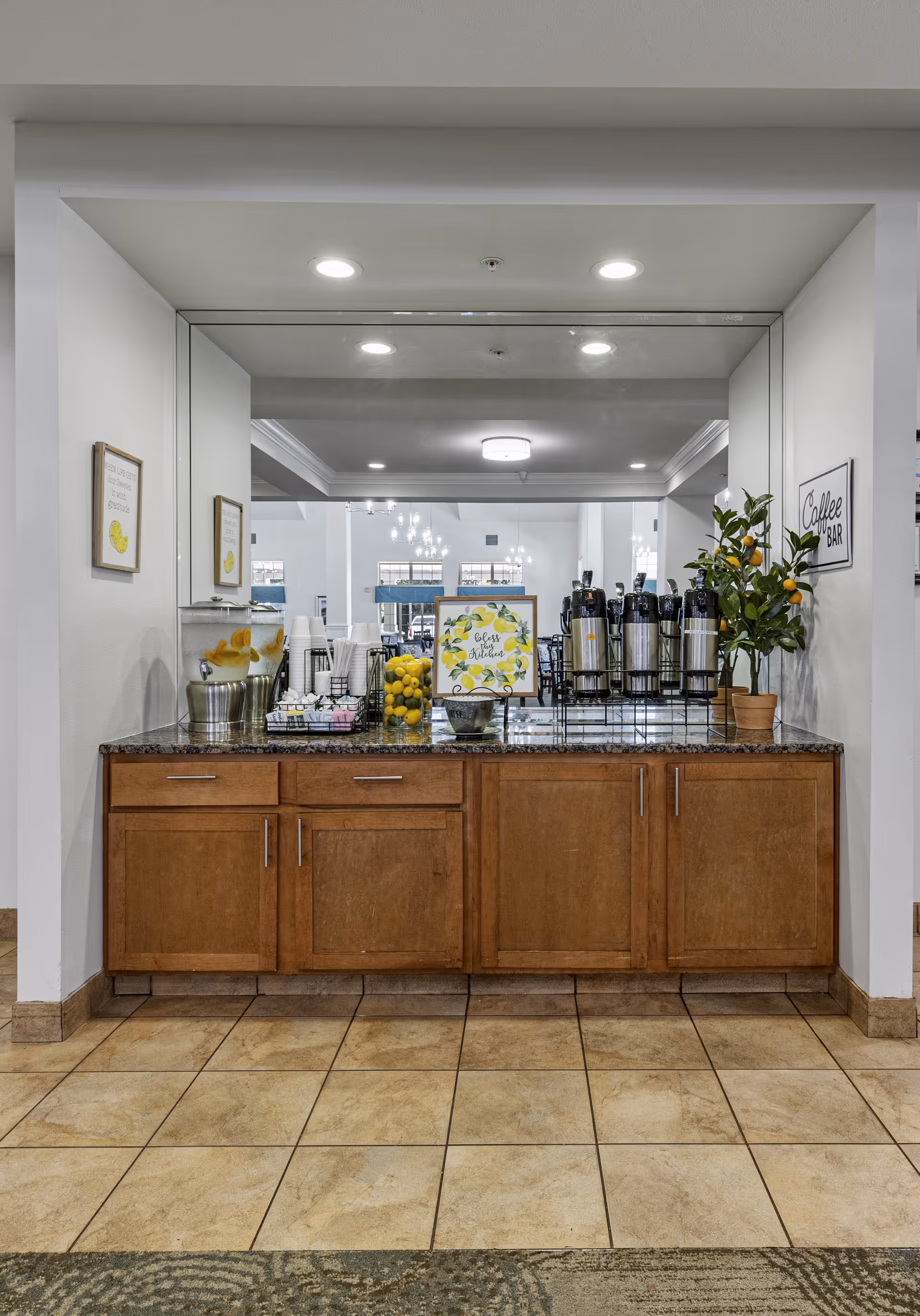 A coffee bar area in a senior living facility with a granite countertop, wooden cabinets, and a large mirror behind. The counter holds several coffee dispensers, a water dispenser with lemon slices, cups, a bowl of lemons, a small potted plant, and a decorative sign that says 'Coffee & Kindness'. The floor is tiled and the walls are white with framed pictures and a 'Coffee Bar' sign.