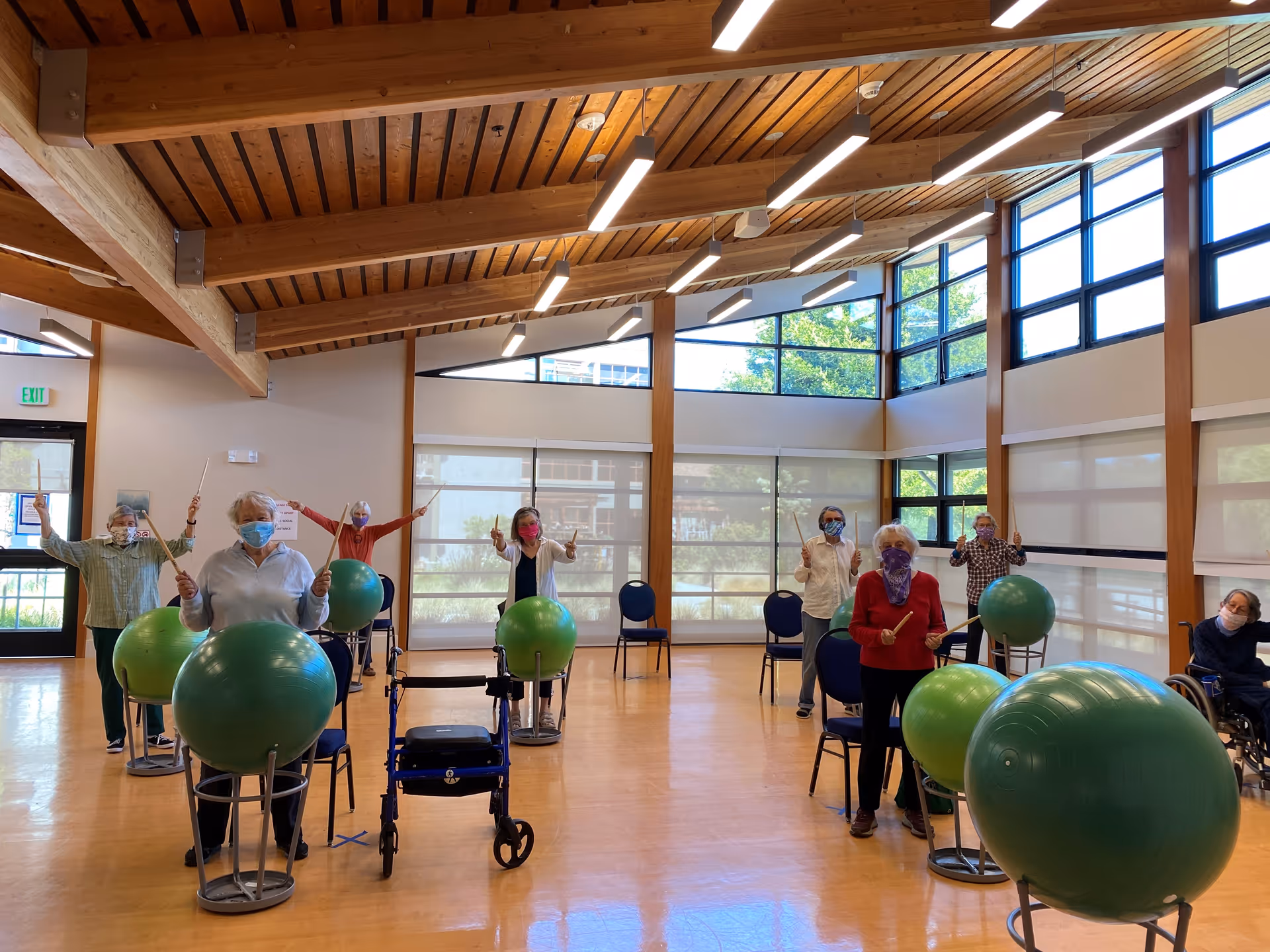 A group of elderly individuals wearing masks participate in a seated exercise class using large green exercise balls and drumsticks in a spacious room with wooden beams and large windows.