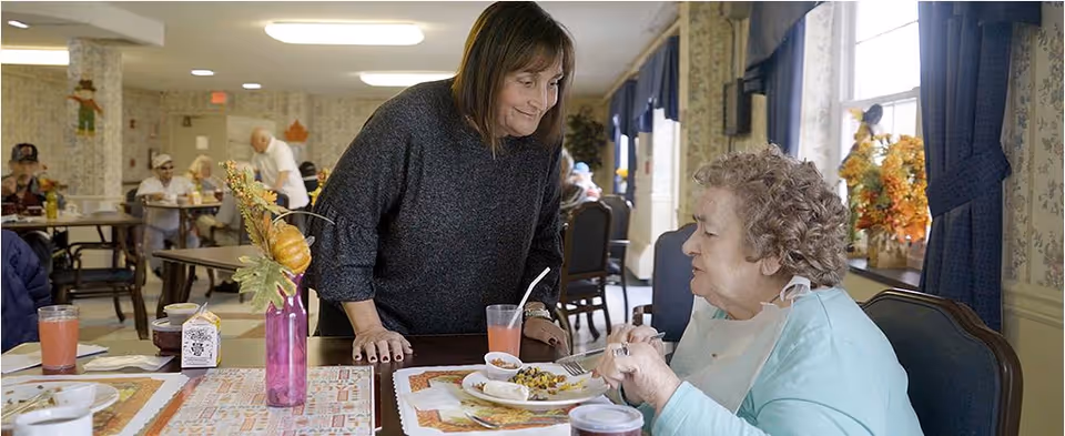 A woman in a dark sweater leans over to talk to an elderly woman seated at a dining table in a nursing facility dining room. The elderly woman is wearing a light blue top and a bib, eating a meal with a fork. The table has placemats, drinks, and a small vase with autumn-themed decorations. Other elderly residents are visible in the background seated at tables.