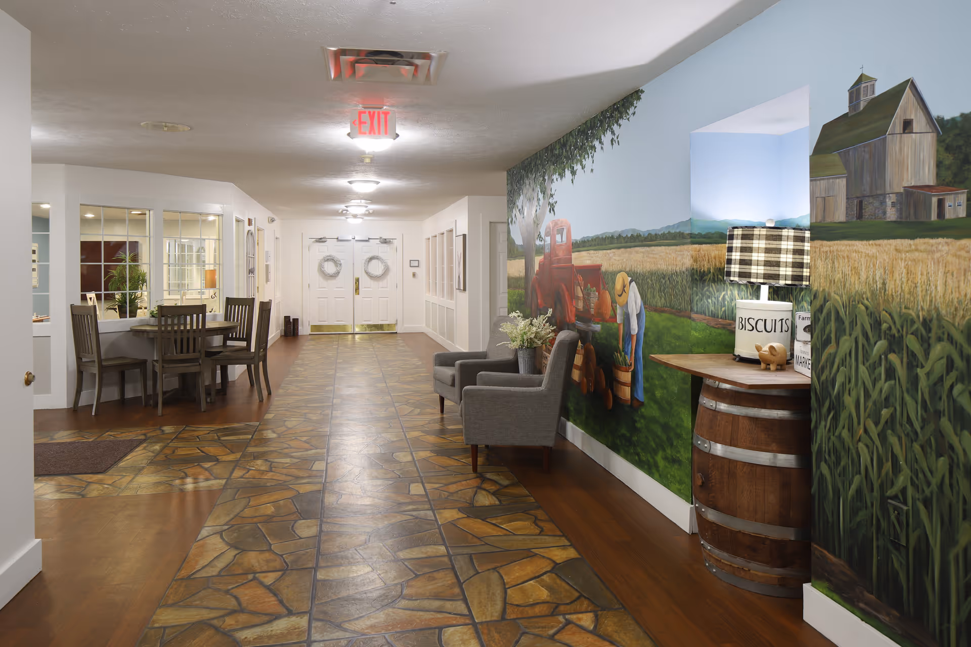 A hallway in Buchanan Meadows facility with a stone-patterned floor and white walls. On the right side, there is a mural depicting a farm scene with a barn, a red tractor, and a person working in a field. Two gray armchairs and a wooden barrel table with a lamp and decorative items are placed against the mural wall. On the left side, there is a dining area with a round table and chairs, and double doors with wreaths at the end of the hallway.
