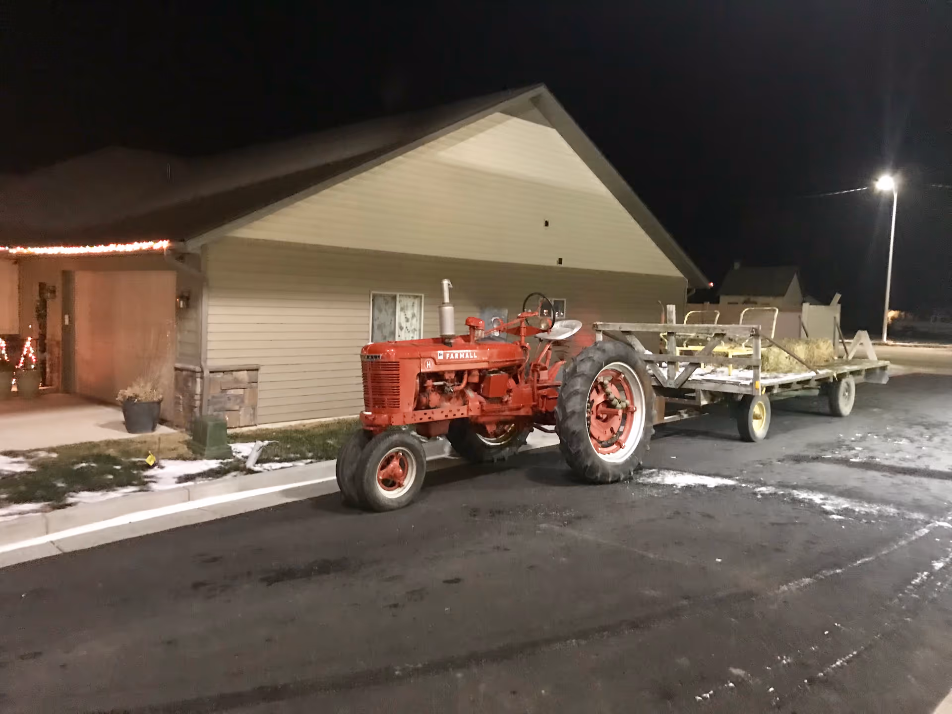 A red tractor attached to a trailer filled with hay is parked on a paved area outside a beige building at night. The building has a sloped roof and some holiday lights near the entrance. There is a streetlight illuminating the scene and patches of snow on the ground.