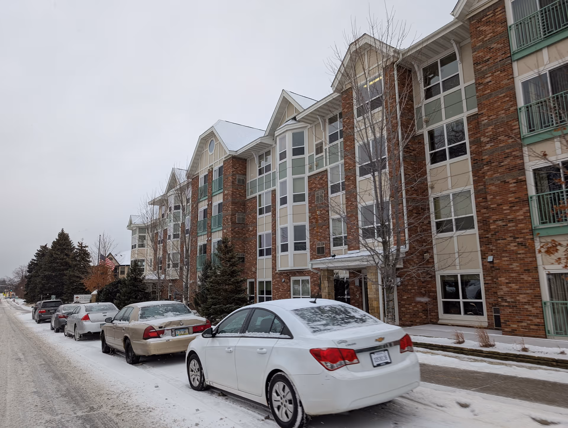 Exterior view of Valora Senior Living of St. Anthony, a multi-story brick and beige building with large windows and green balconies. Several cars are parked along a snow-covered street in front of the building, with leafless trees and an American flag visible.