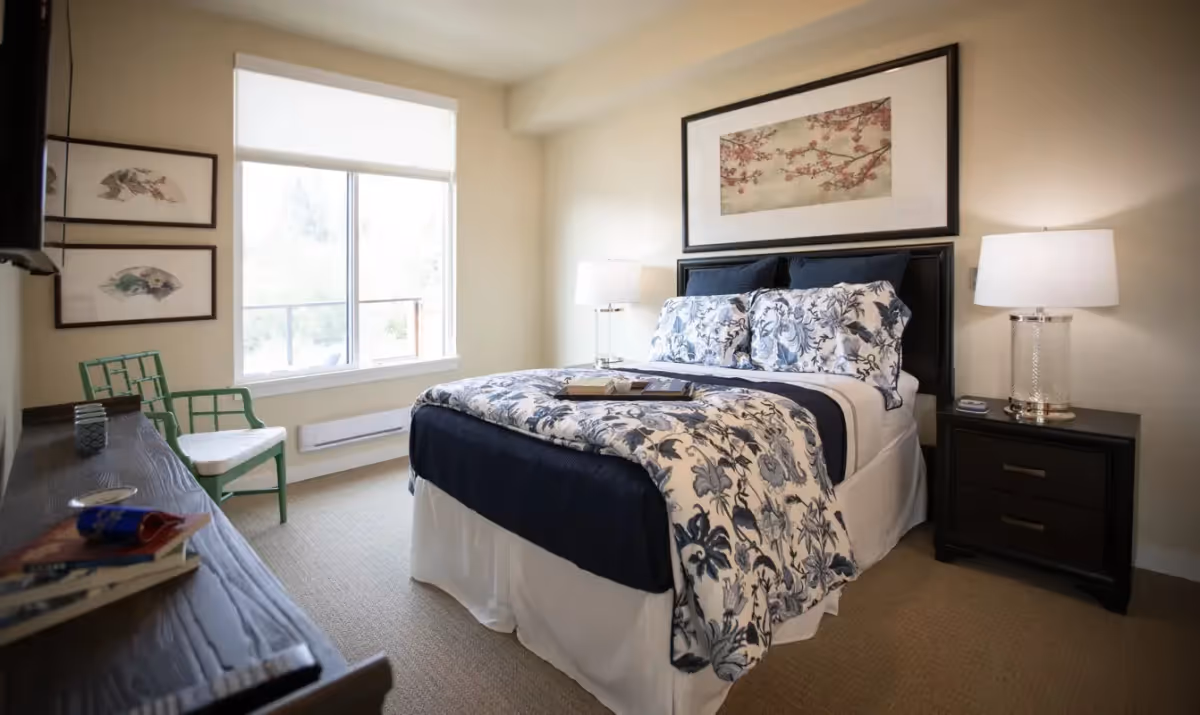 A cozy bedroom with a neatly made bed featuring floral patterned bedding in blue and white. The room has a large window with a white blind, a green chair, a dark wooden dresser with books and items on top, two framed pictures on the wall, and a nightstand with a lamp beside the bed. The walls are painted a light beige color.