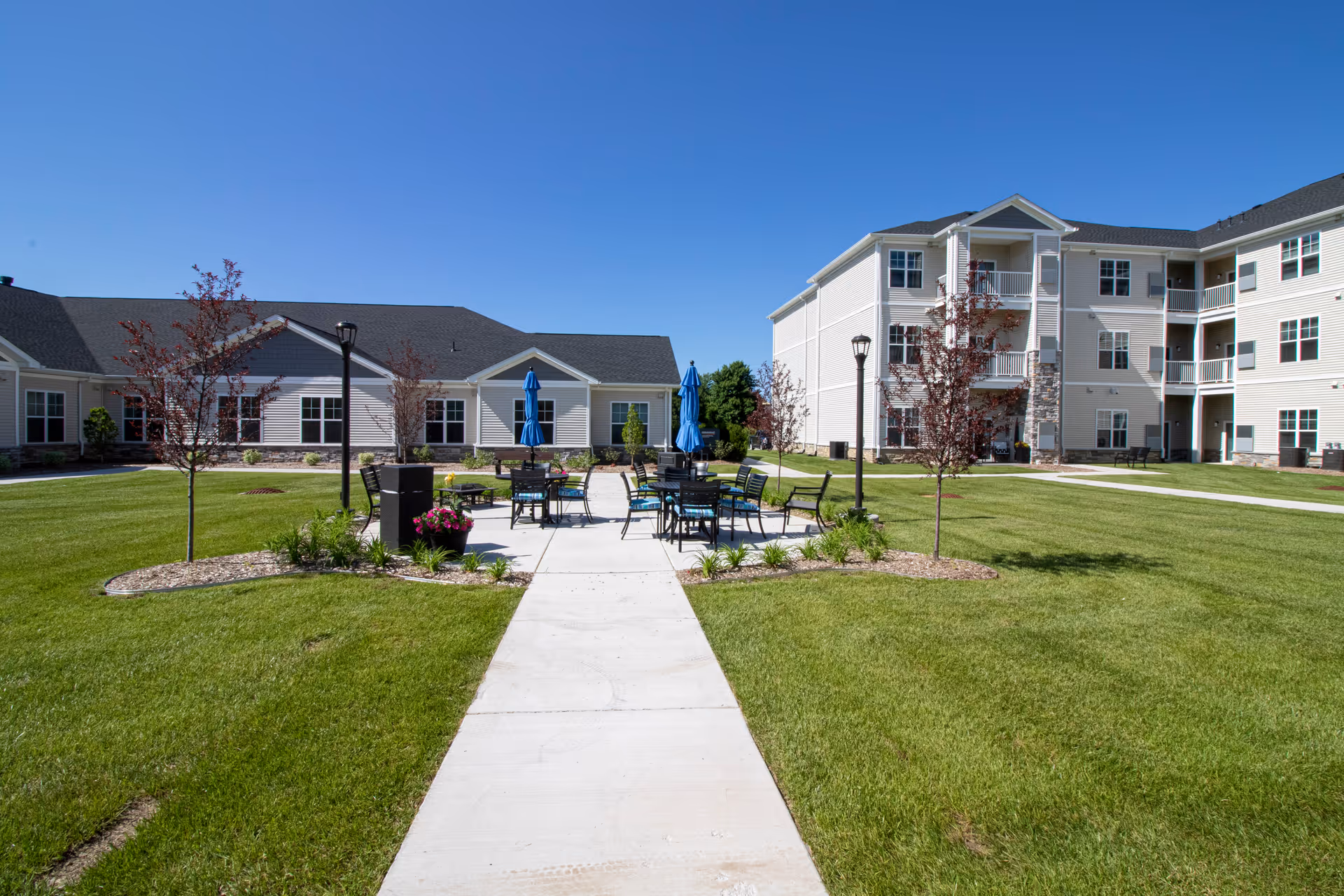 Courtyard with patio tables and umbrellas between multi-story senior living buildings under a clear blue sky.