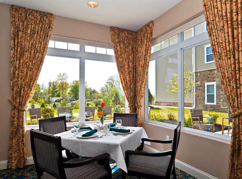 A dining area with a square table covered with a white tablecloth, set with plates, glasses, silverware, and green napkins. Four dark wooden chairs with cushions surround the table. Large windows with floral curtains provide a view of an outdoor patio with chairs and greenery.