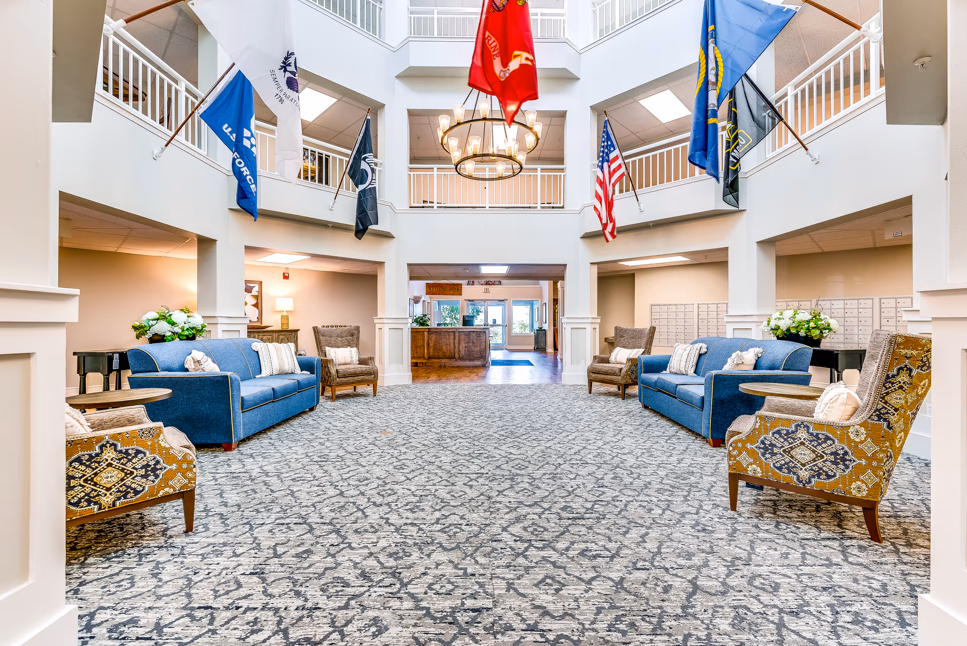 Spacious two-story senior living lobby with seating areas, patterned carpet, flags hanging from the upper balcony, and a central chandelier.