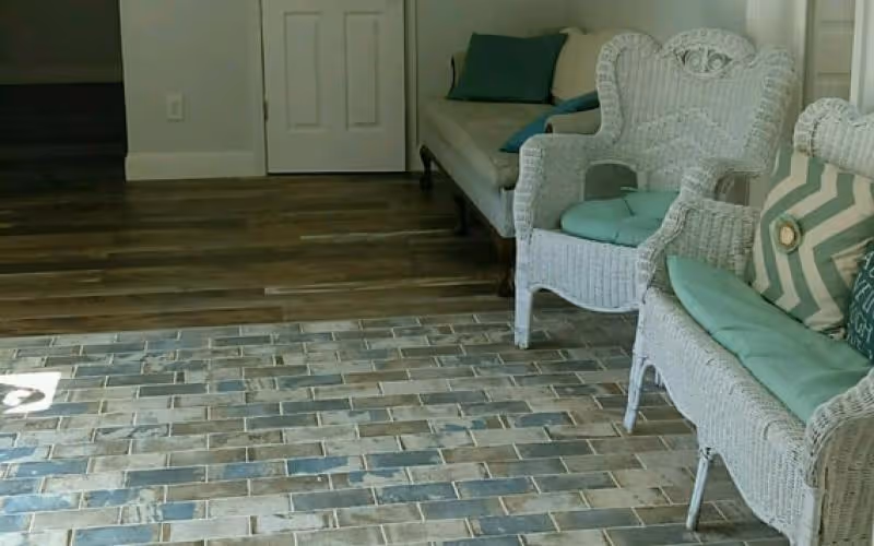 Interior seating area with two white wicker chairs with light blue cushions and a matching loveseat with green and patterned pillows, set on a floor with a mix of wooden and brick-like tiles. A closed white door and a staircase are visible in the background.
