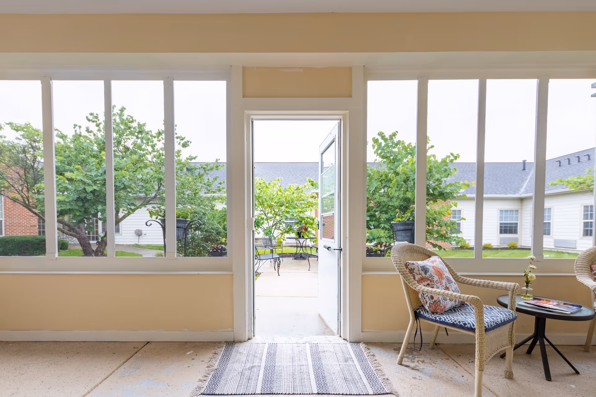 A bright indoor seating area with large windows looking out onto a garden courtyard. There are two wicker chairs with patterned cushions and a small round table with a flower vase and magazines. The door in the center is open, leading outside to a patio with metal chairs and tables surrounded by greenery and trees.