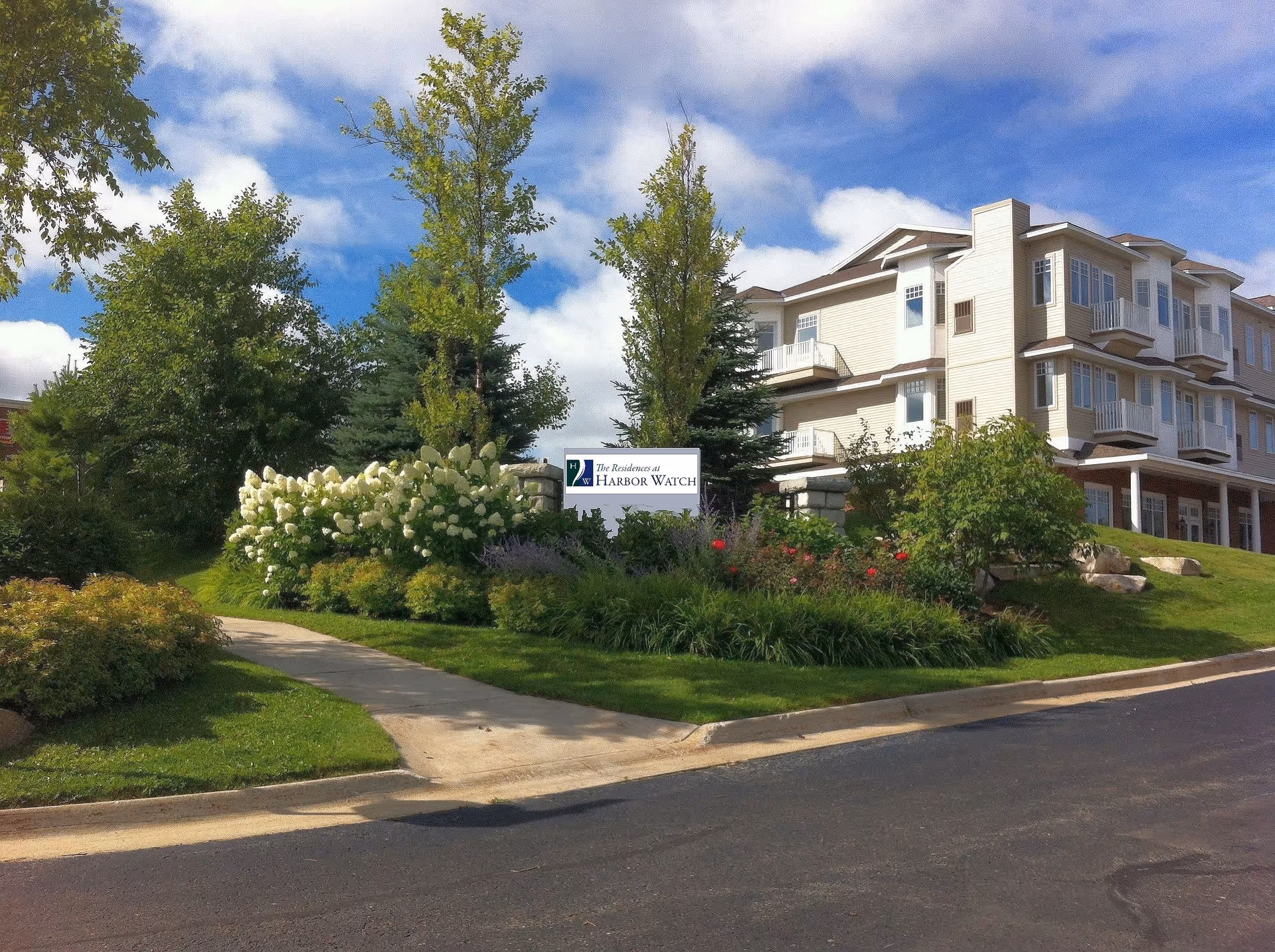 Exterior view of The Residences at Harbor Watch senior living facility showing a multi-story building with balconies surrounded by landscaped greenery, trees, and flowering bushes under a partly cloudy sky.
