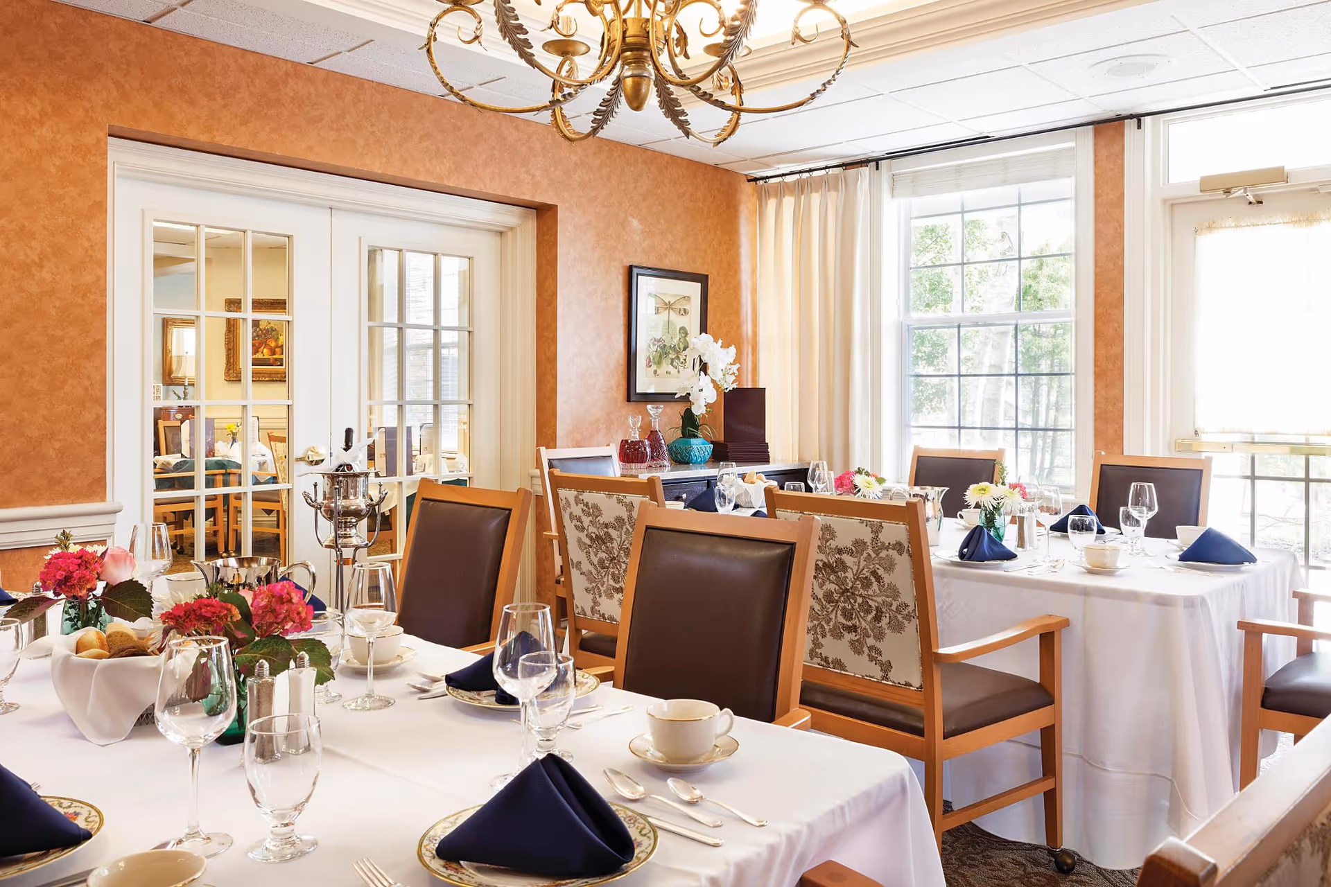 Formal dining room with tables set with white tablecloths, glassware, navy napkins, floral centerpieces, and wooden chairs by large windows.
