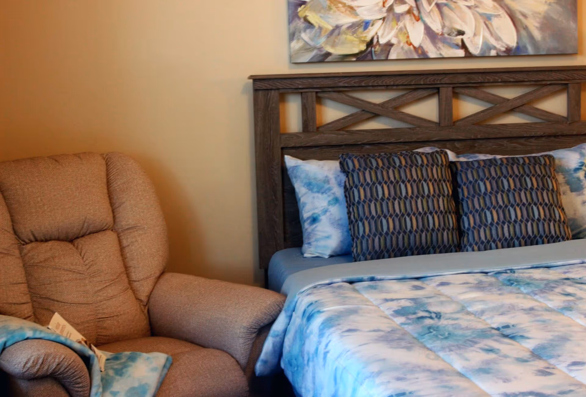 A cozy bedroom featuring a wooden headboard bed with blue and white patterned bedding and pillows, next to a comfortable beige recliner chair with a blue blanket draped over the arm.