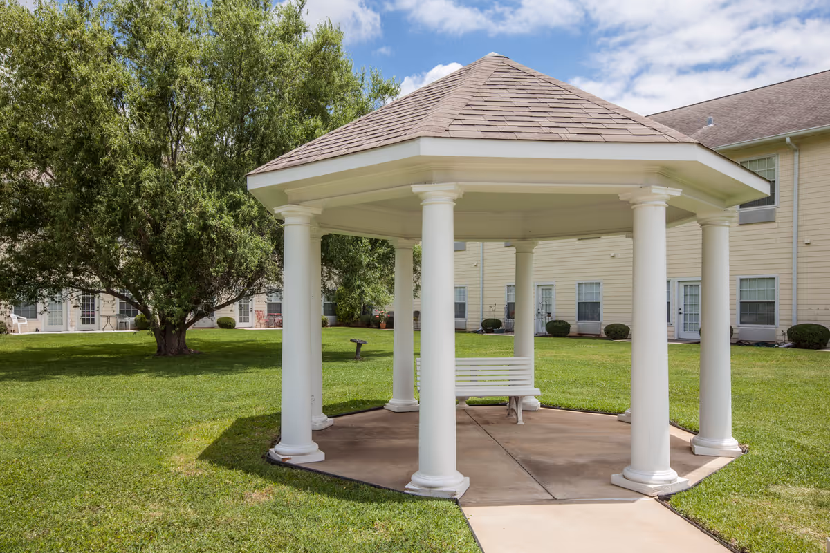 White gazebo with columns and a bench on a grassy lawn in front of a residential building.