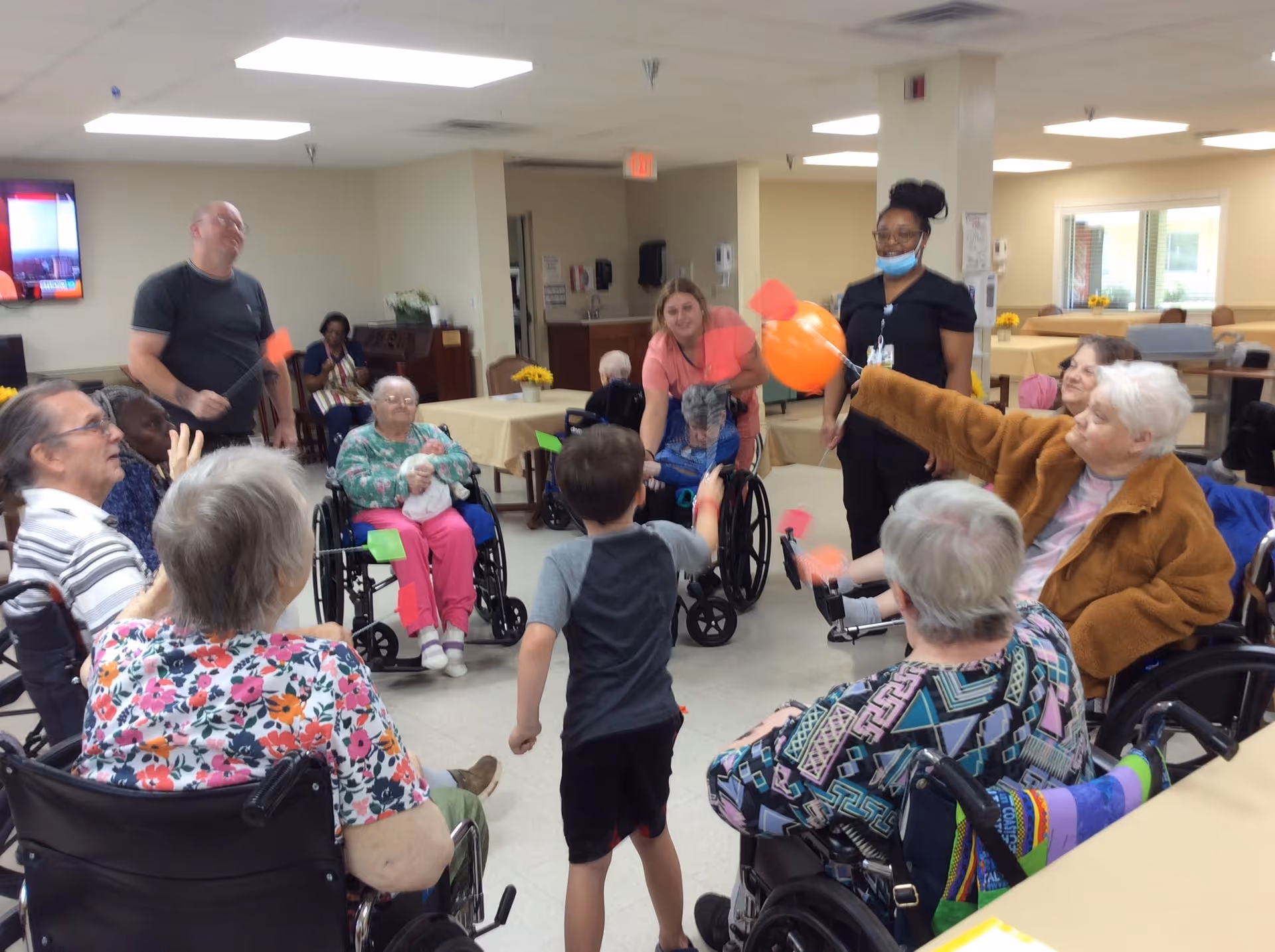 A group of elderly people, some in wheelchairs, and a few caregivers and a child are gathered in a common room of a senior living facility. They are engaged in a group activity involving orange balloons and small paddles. The room has tables with yellow flowers and a television mounted on the wall.