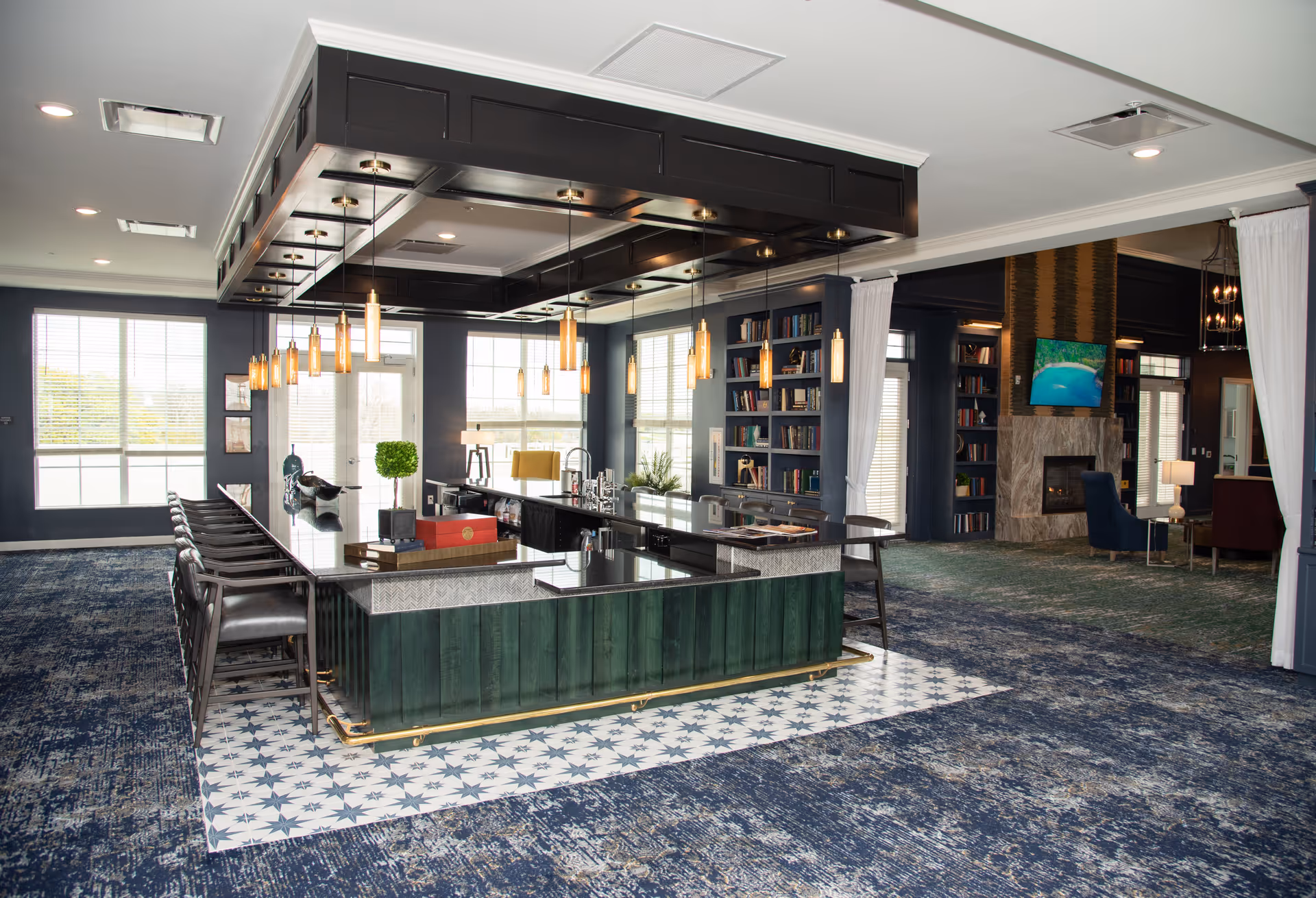 Spacious senior living common area with a central bar island surrounded by stools, pendant lights, bookshelves and a seating area with a fireplace.
