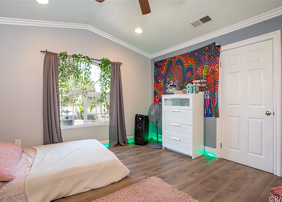 A bedroom with a bed covered in light-colored bedding and a pink pillow. There is a window with gray curtains and green hanging plants. A white dresser stands against the wall with a colorful abstract tapestry hanging above it. A standing fan and an air purifier are placed next to the dresser. The room has wood flooring and a white door.