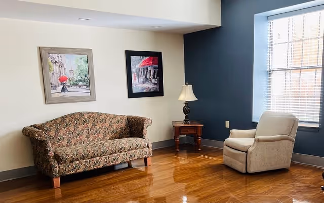 A cozy sitting area in a living room with a floral patterned sofa, a beige armchair, a wooden side table with a lamp, two framed paintings on the wall, and a large window with blinds letting in natural light.