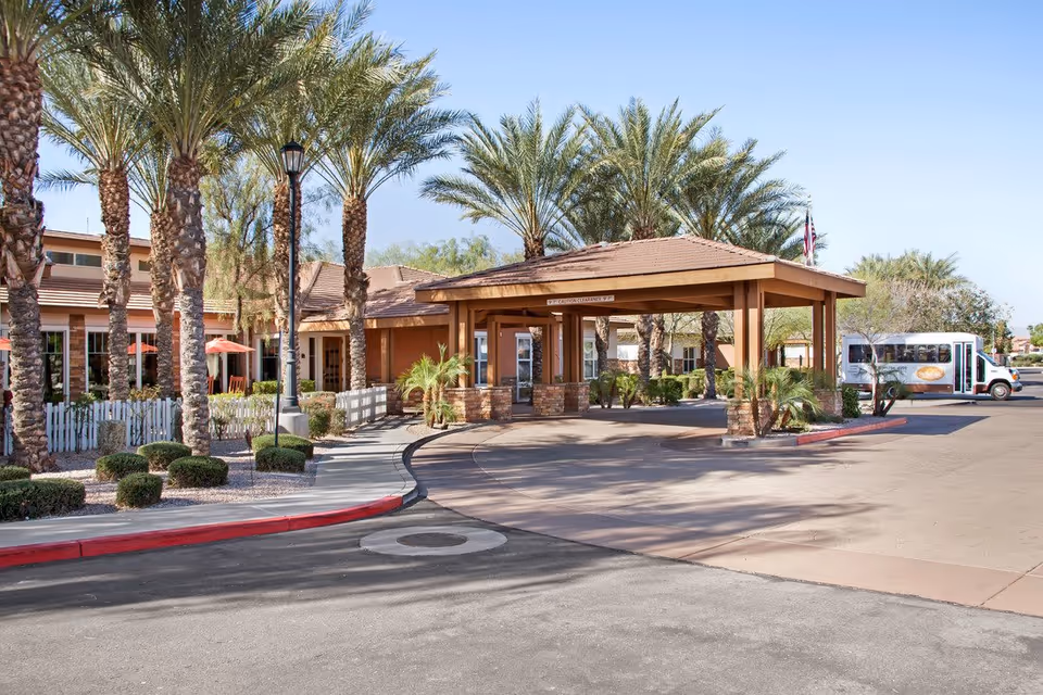 Exterior view of Sunrise of Chandler senior living facility entrance with a covered drop-off area, palm trees, a white picket fence, and a shuttle bus parked nearby under a clear blue sky.