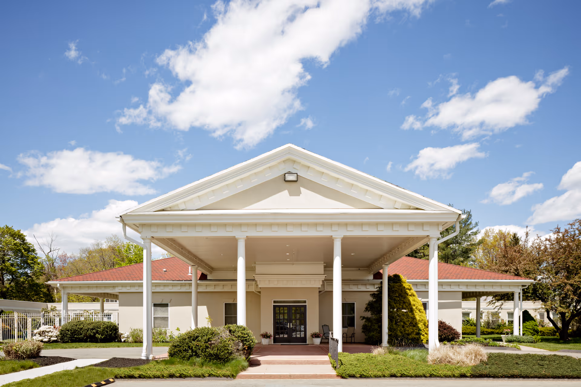 Front exterior view of Autumn Lake Healthcare at Bucks Hill building with a covered entrance supported by white columns, surrounded by greenery and under a partly cloudy blue sky.
