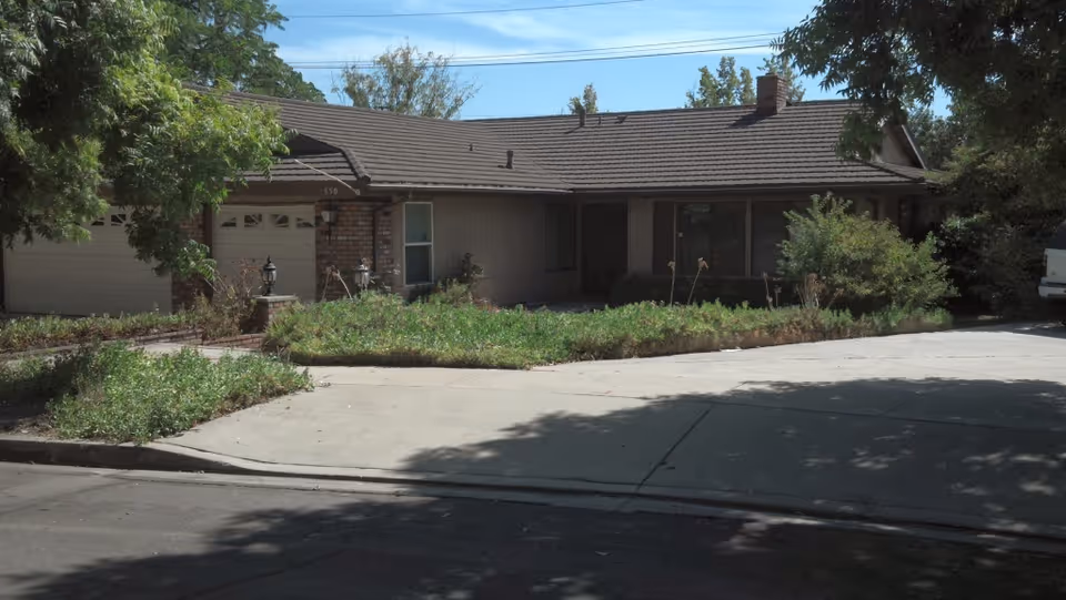 Front exterior of a single-story ranch-style house with a driveway, garage, and overgrown landscaping.