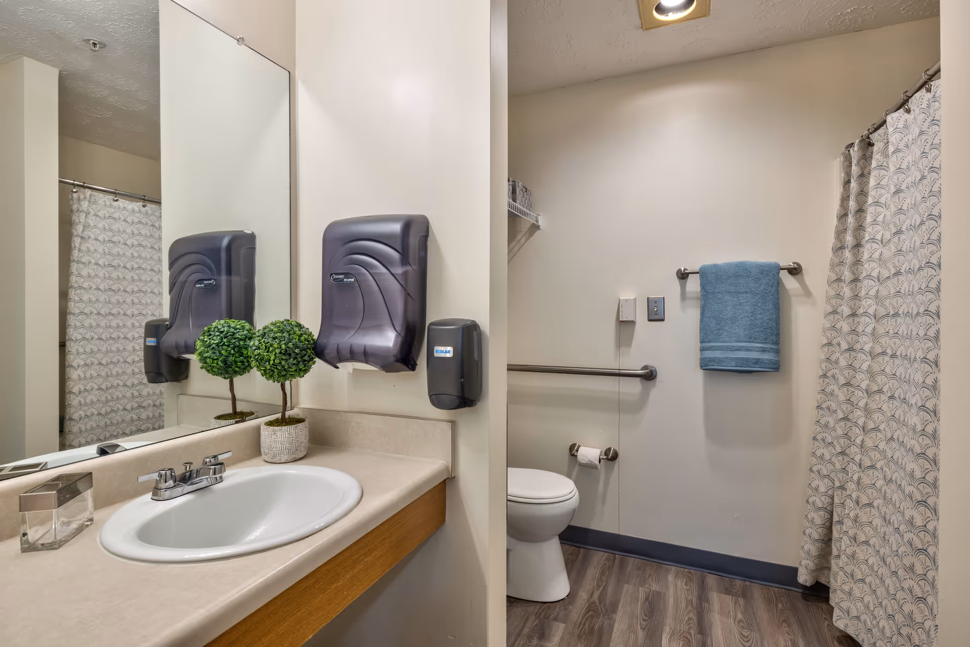 A clean bathroom with a sink and countertop on the left side, featuring a small decorative plant and a soap dispenser. Above the sink is a large mirror with a paper towel dispenser and a hand sanitizer dispenser mounted on the wall. On the right side, there is a toilet with a grab bar beside it, a toilet paper holder, a towel rack with a blue towel, and a shower area with a patterned shower curtain. The floor has wood-like vinyl flooring.