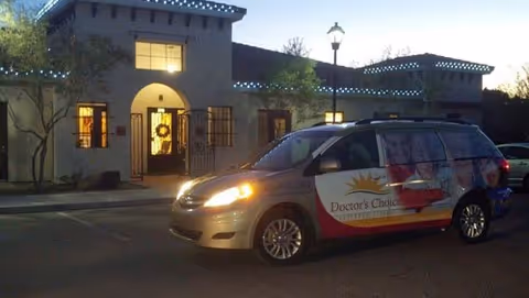 Exterior view of a building at dusk with lights on inside. A minivan with Doctor's Choice Assisted Living branding is parked in front of the building.