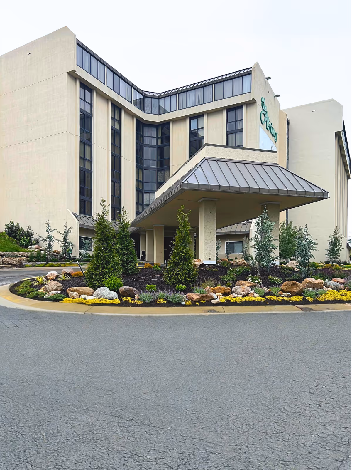 Exterior view of The Atriums Senior Living Community building featuring a multi-story structure with large windows and a covered entrance. The landscaped area in front includes various shrubs, small trees, rocks, and yellow flowering plants.