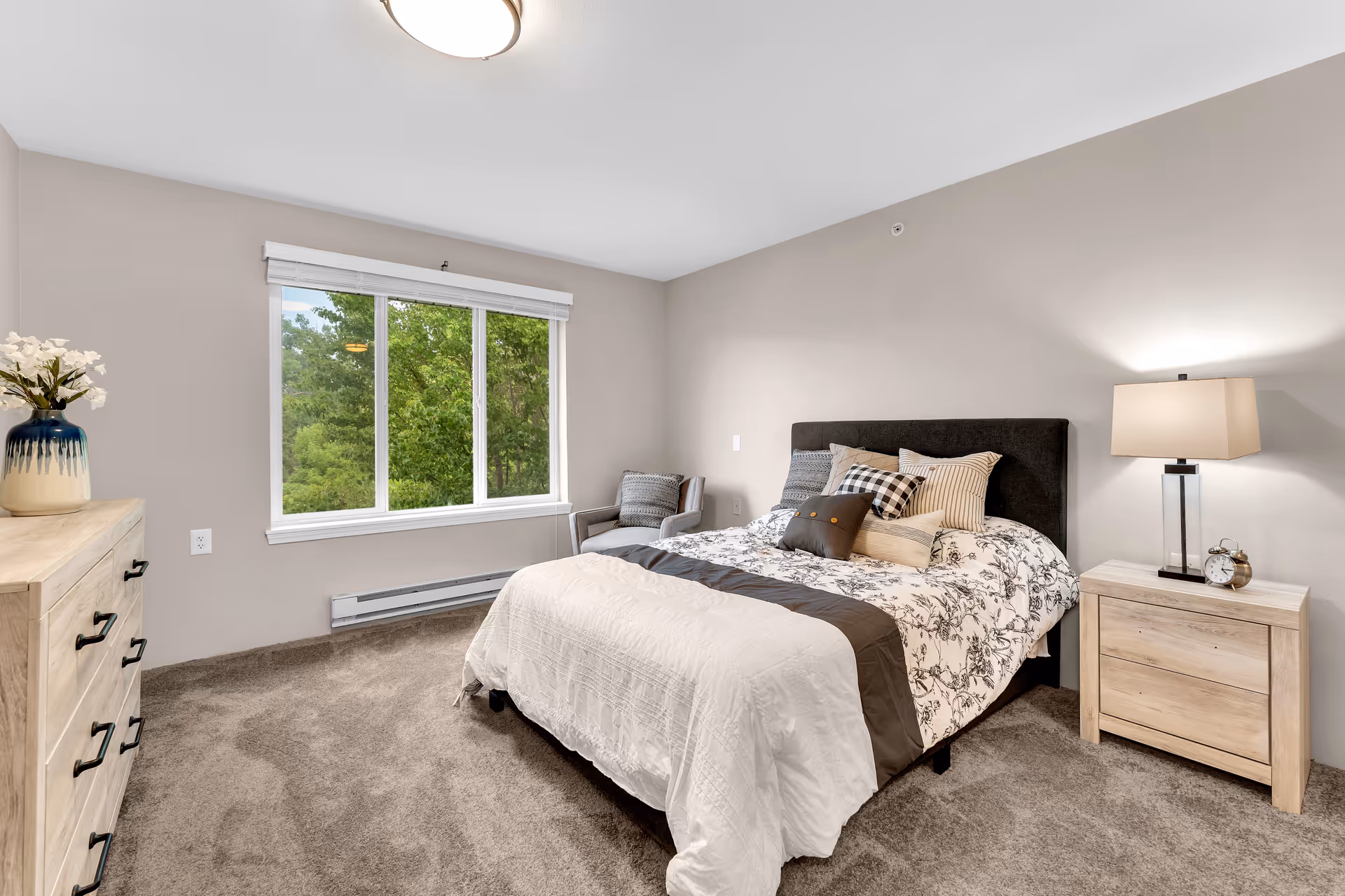 A cozy bedroom with a large window showing green trees outside. The room features a bed with a black headboard, white and patterned bedding, and multiple pillows. There is a wooden nightstand with a lamp and an alarm clock on the right side of the bed, and a wooden dresser with a vase of white flowers on the left side. A cushioned chair is placed near the window. The walls are painted light beige and the floor is carpeted.