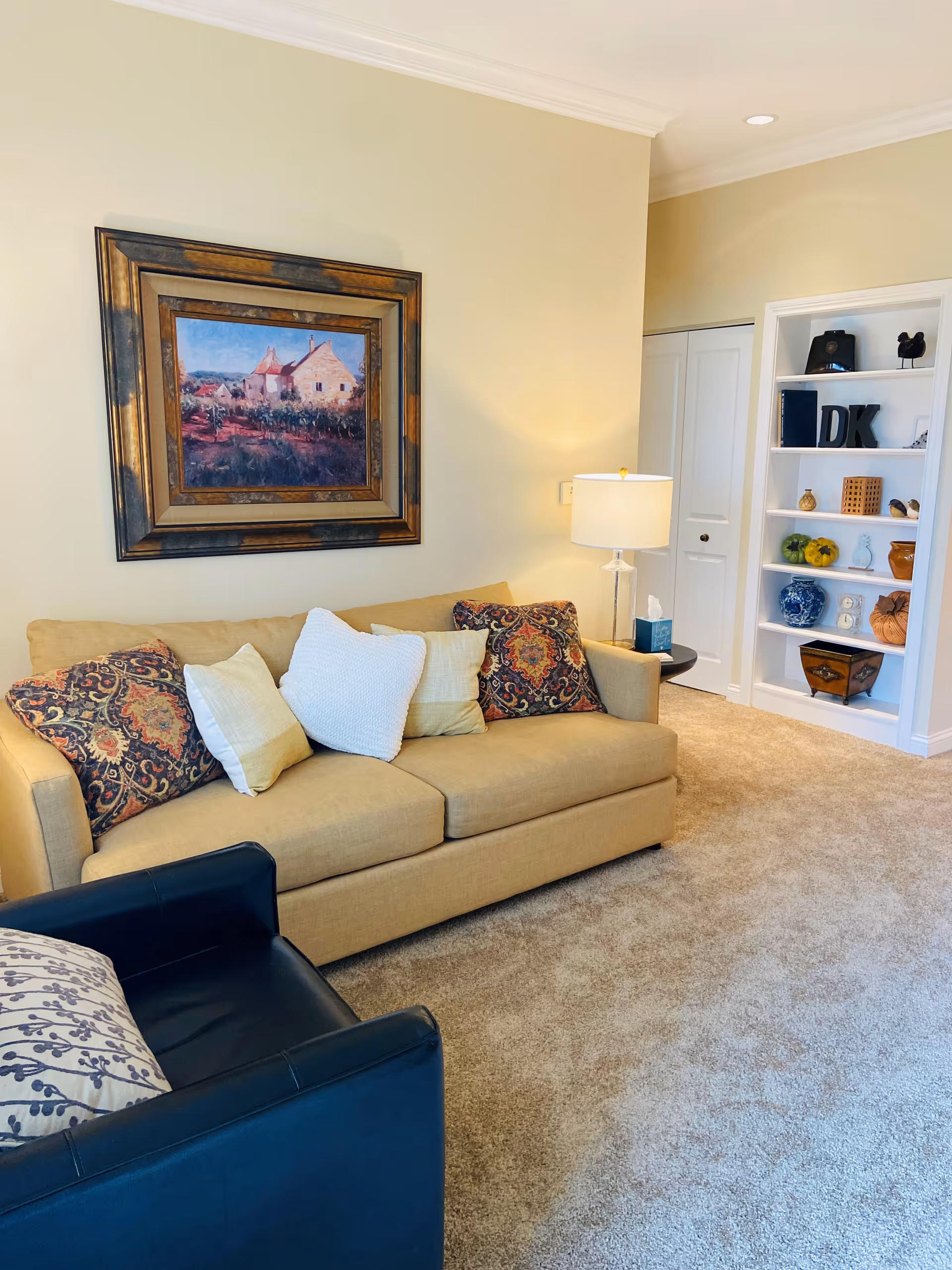 A cozy living room with a beige sofa adorned with patterned and plain cushions, a black armchair with a decorative pillow, a floor lamp with a white shade, a framed painting of a house on the wall, and a built-in white bookshelf displaying various decorative items.