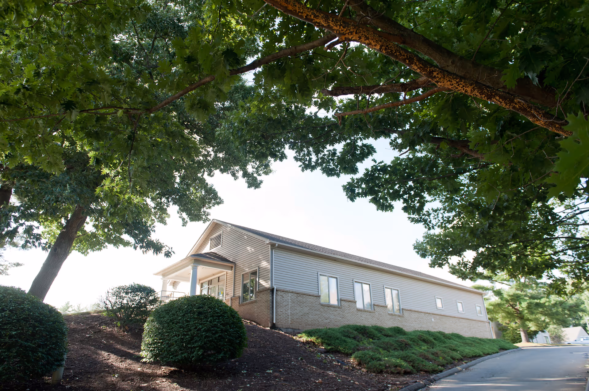 Exterior view of a single-story building with beige siding and brick foundation, surrounded by green bushes and large trees with dense foliage, under a clear sky.