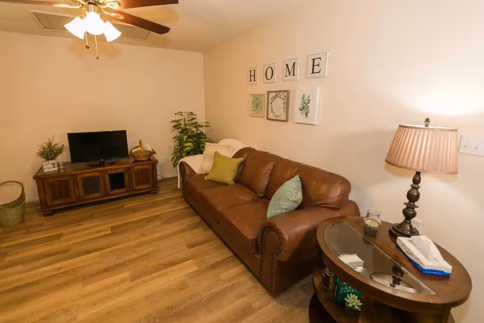 A cozy living room with a brown leather sofa adorned with green and blue pillows, a wooden TV stand with a flat-screen TV, a ceiling fan with lights, a side table with a lamp and tissue box, and wall decor spelling 'HOME' above framed botanical prints.