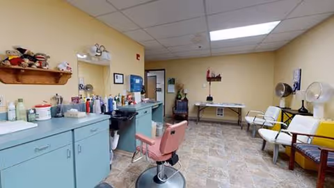 Interior salon room with teal styling stations and mirrors, a pink barber chair, several waiting chairs and a table in a beige-tiled room.