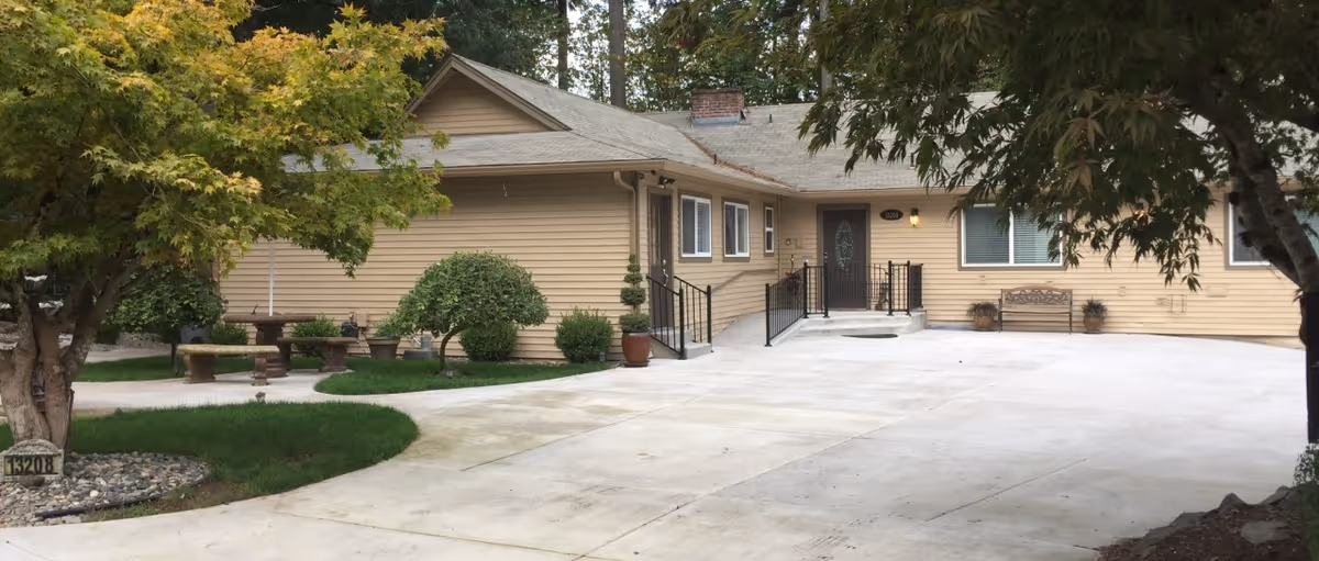Single-story beige house front with a wide concrete driveway, ramped entrance, bench and landscaped trees and shrubs.