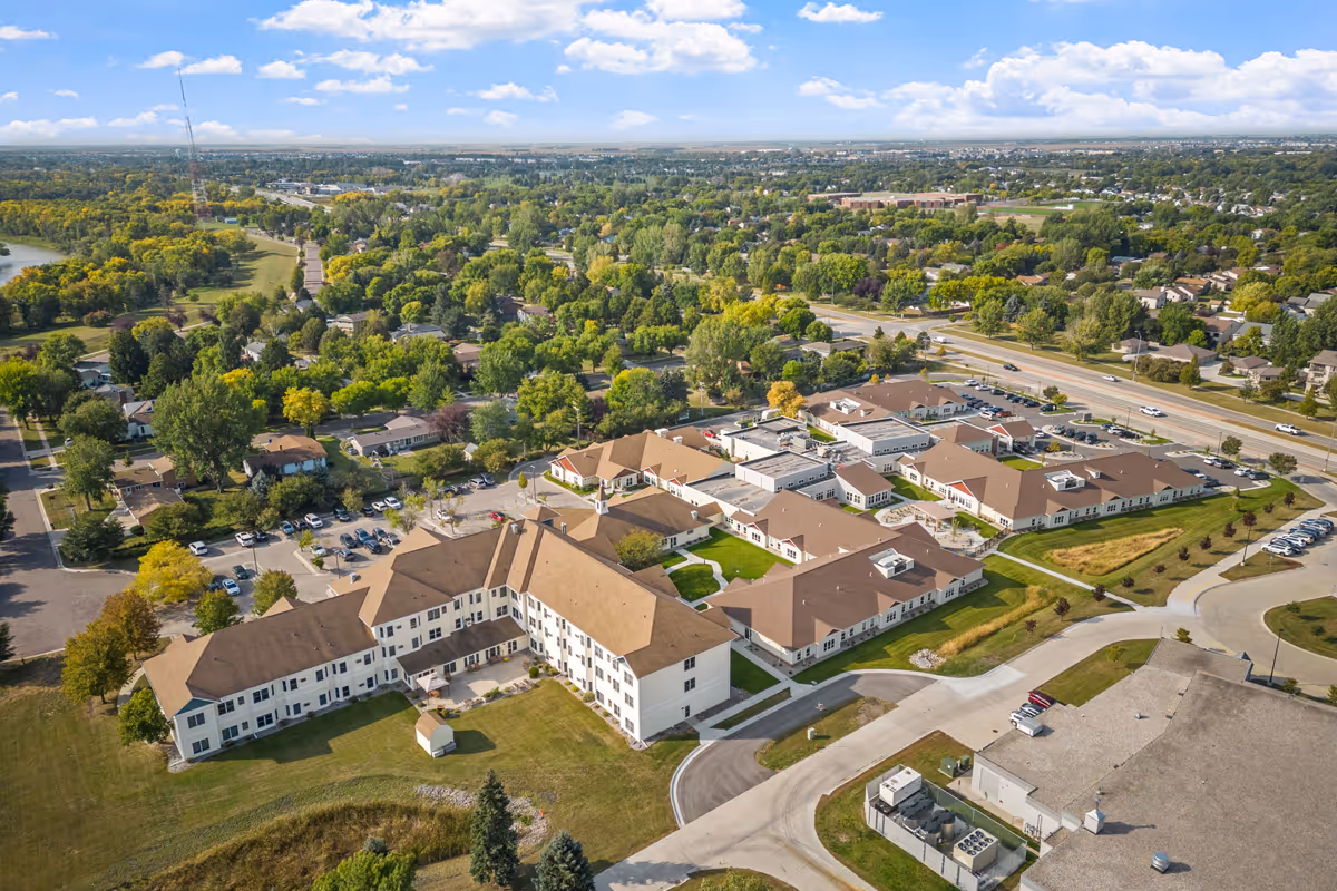 Aerial view of Fargo Elim Senior Living Community showing multiple connected buildings with brown roofs surrounded by green lawns, trees, parking lots, and nearby roads under a partly cloudy sky.