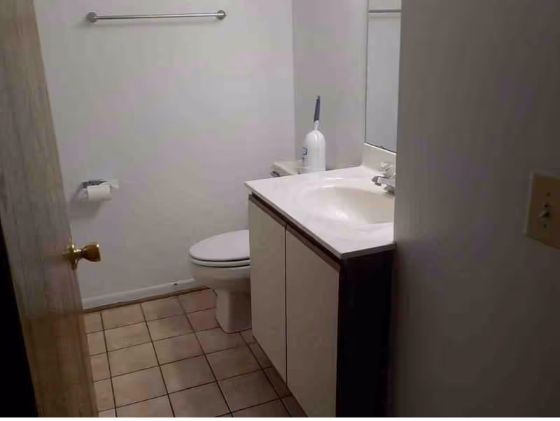 Small bathroom with a toilet, a sink with a cabinet, a mirror above the sink, a towel rack on the wall, and a toilet paper holder. The floor is tiled and the walls are plain white.