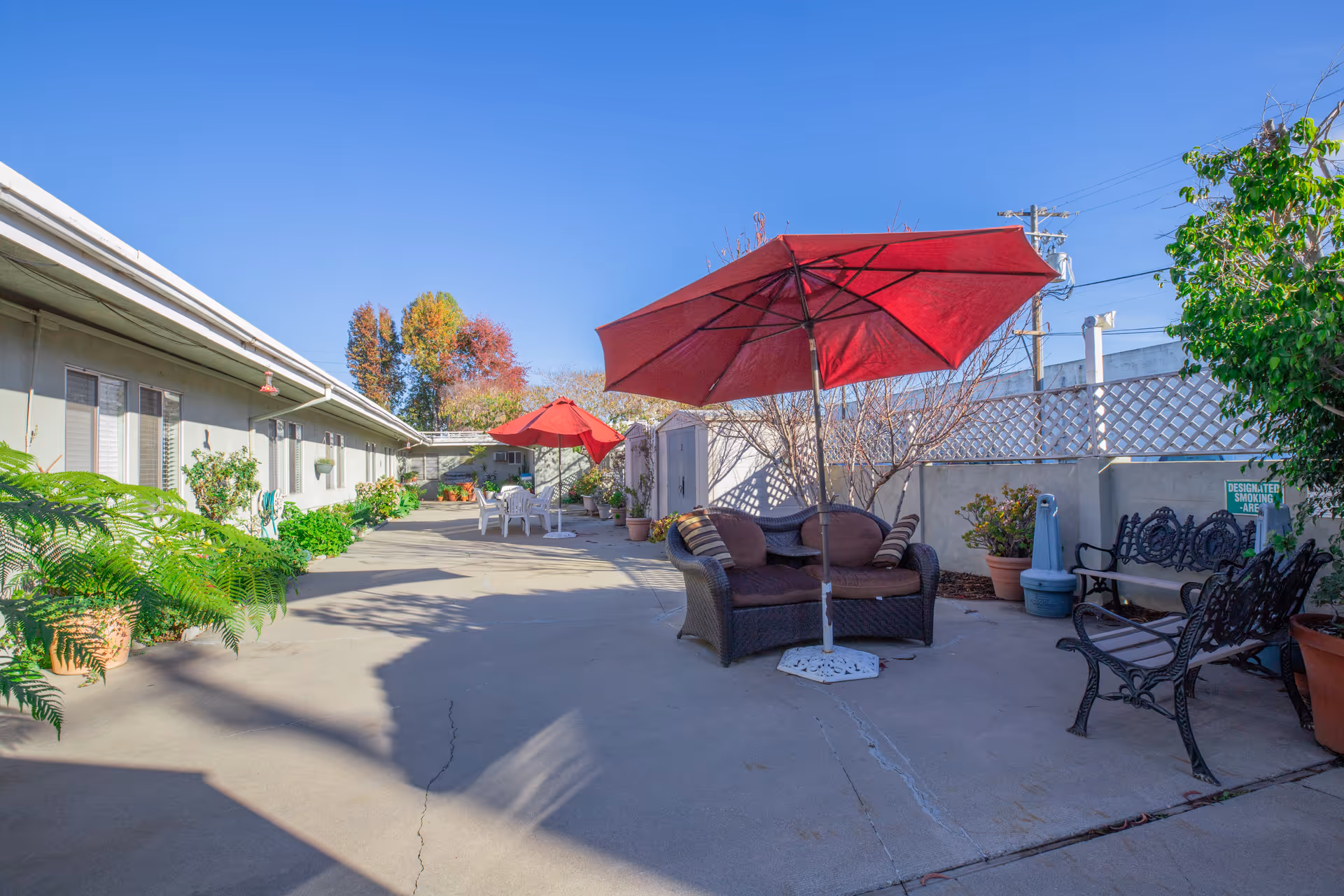 Sunny outdoor courtyard with patio seating, red umbrellas, potted plants, and a walkway beside a low building.